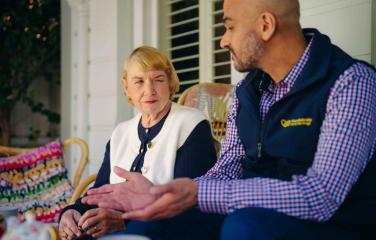 An older lady sits outside with a Vision Australia worker, listening carefully as he explains something to her.